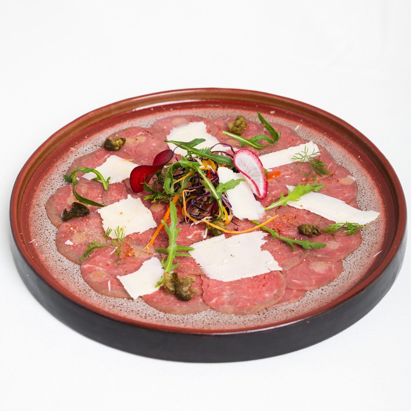 Carpaccio de bœuf dans une assiette rouge, copeaux de parmesan, câpres et roquette