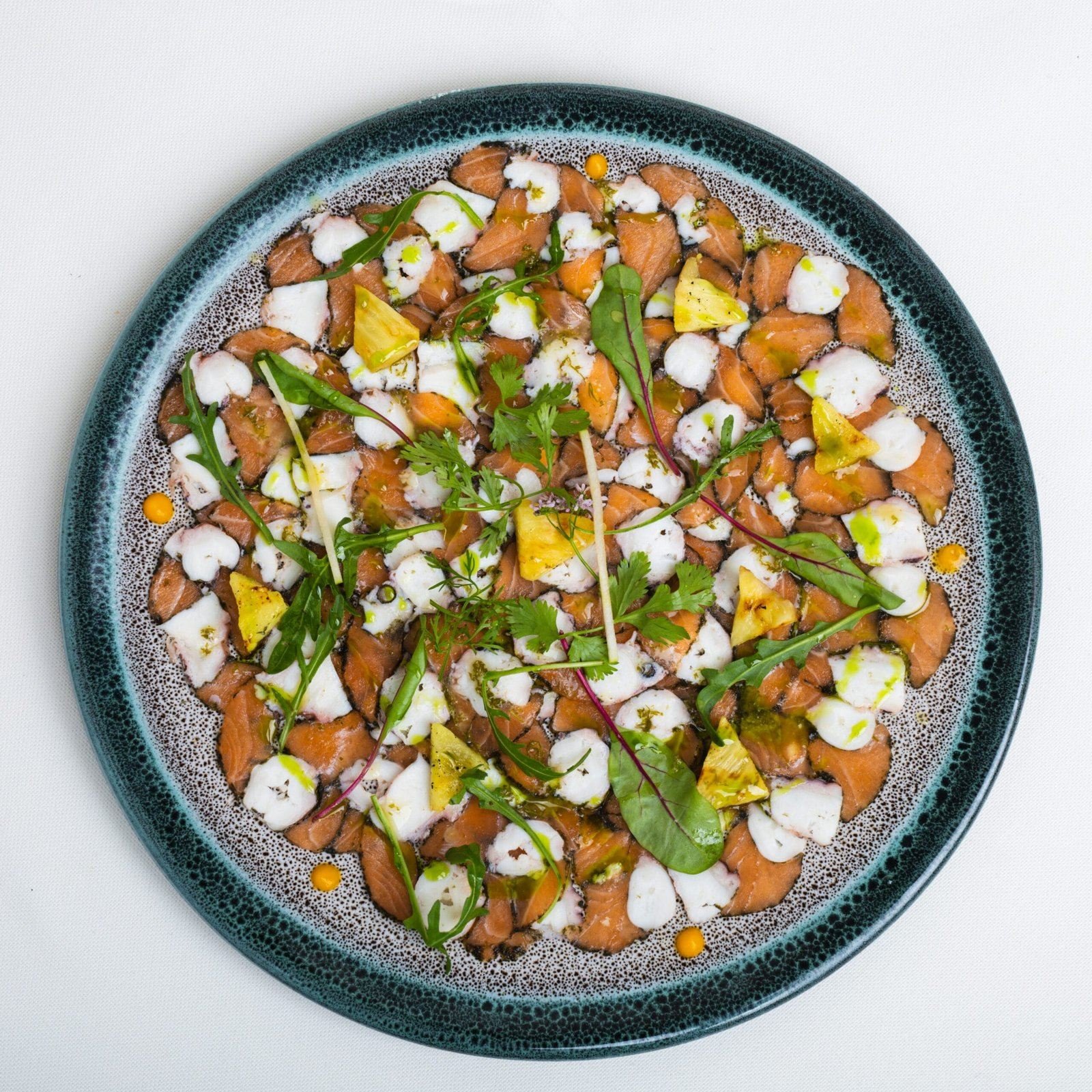 Carpaccio de saumon, ananas rôti, herbes fraîches, dans une assiette bleu nuit