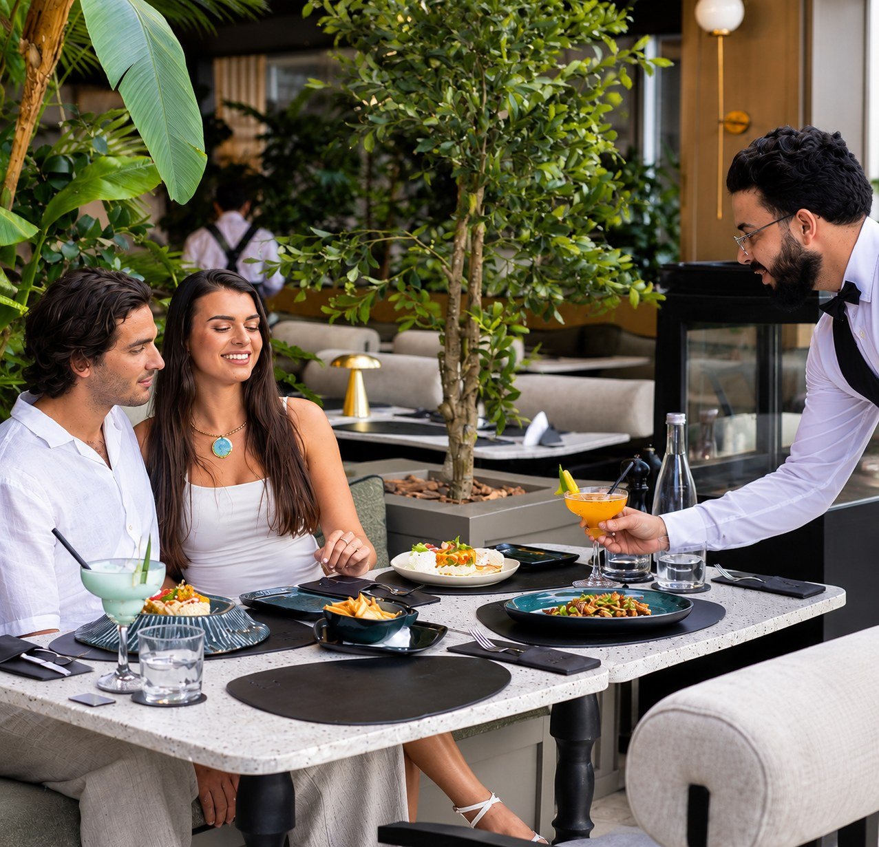 Un couple attablé sur la terrasse, un serveur apporte un cocktail