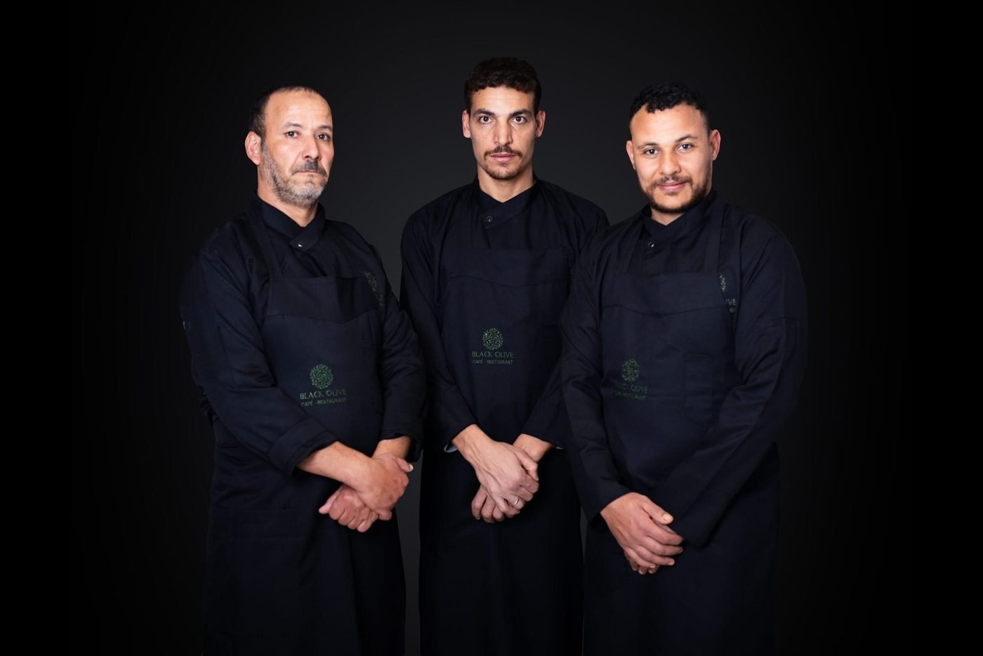 Trois membres de la brigade en tenue noire, tablier Black Olive, sur fond sombre