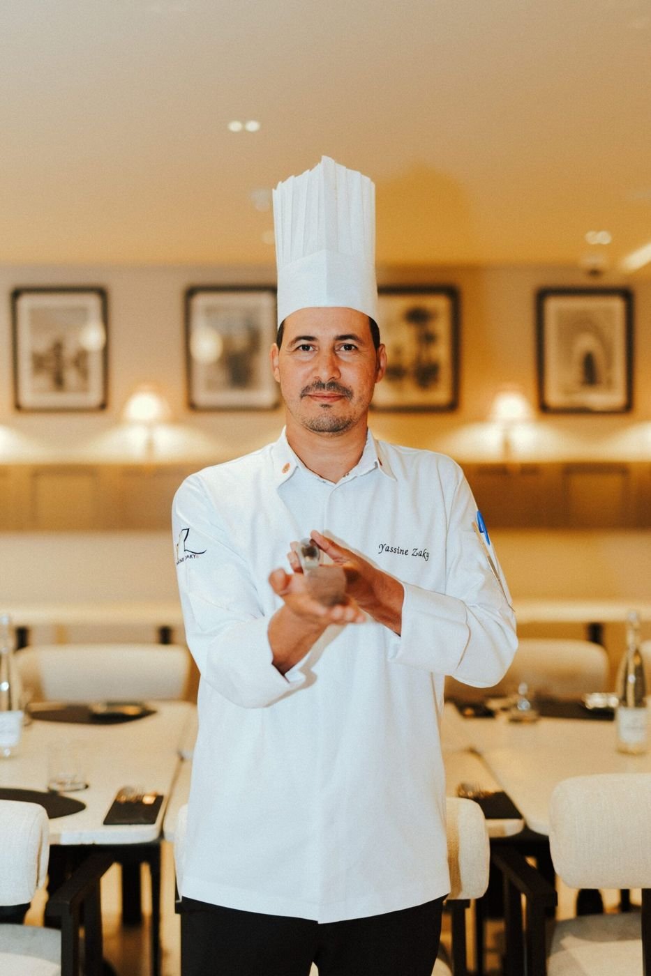 Un chef en tenue blanche, les mains jointes, dans la salle du restaurant