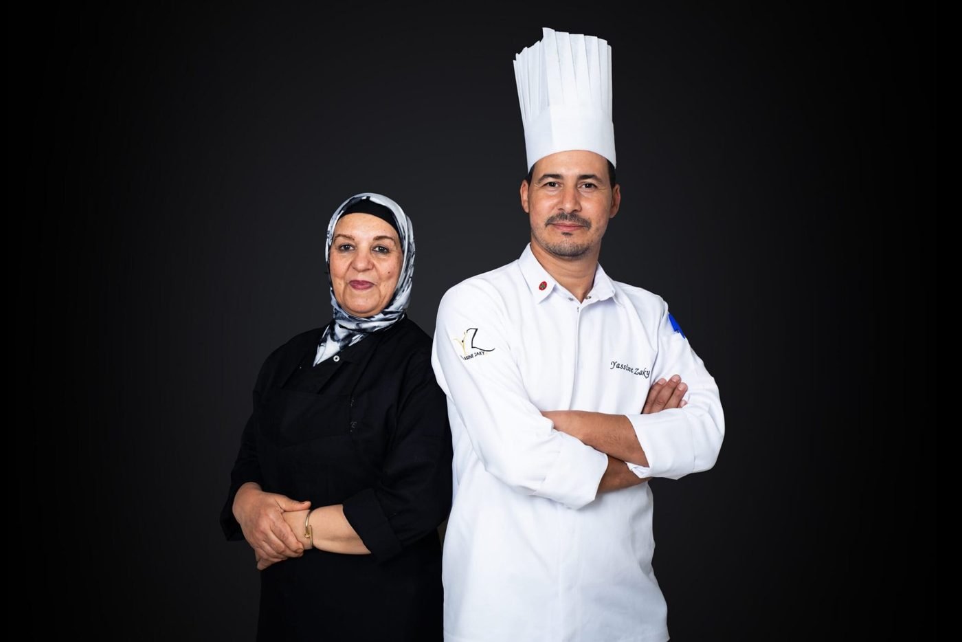 Une cheffe en tenue noire et un chef en tenue blanche, bras croisés, sur fond sombre